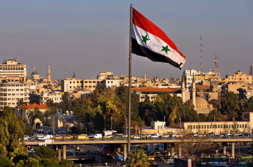 A large Syrian flag is waving in the wind against a backdrop of a city.