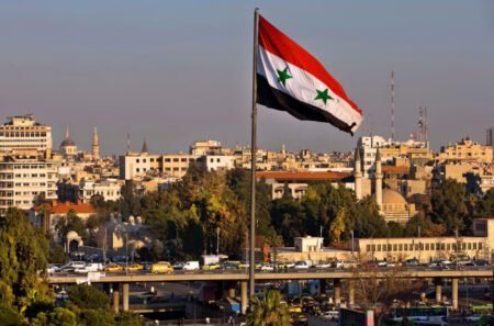 A large Syrian flag is waving in the wind against a backdrop of a city.