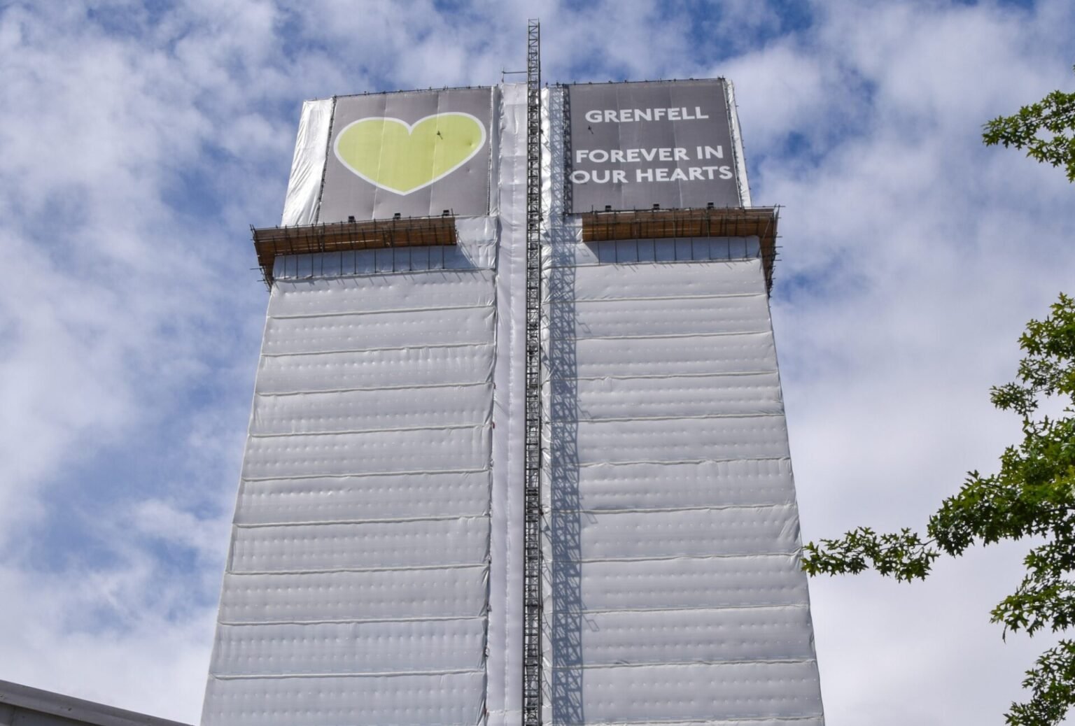 A tall, partially covered building with scaffolding. With a large banner on it which reads "Grenfell Forever in Our Hearts."